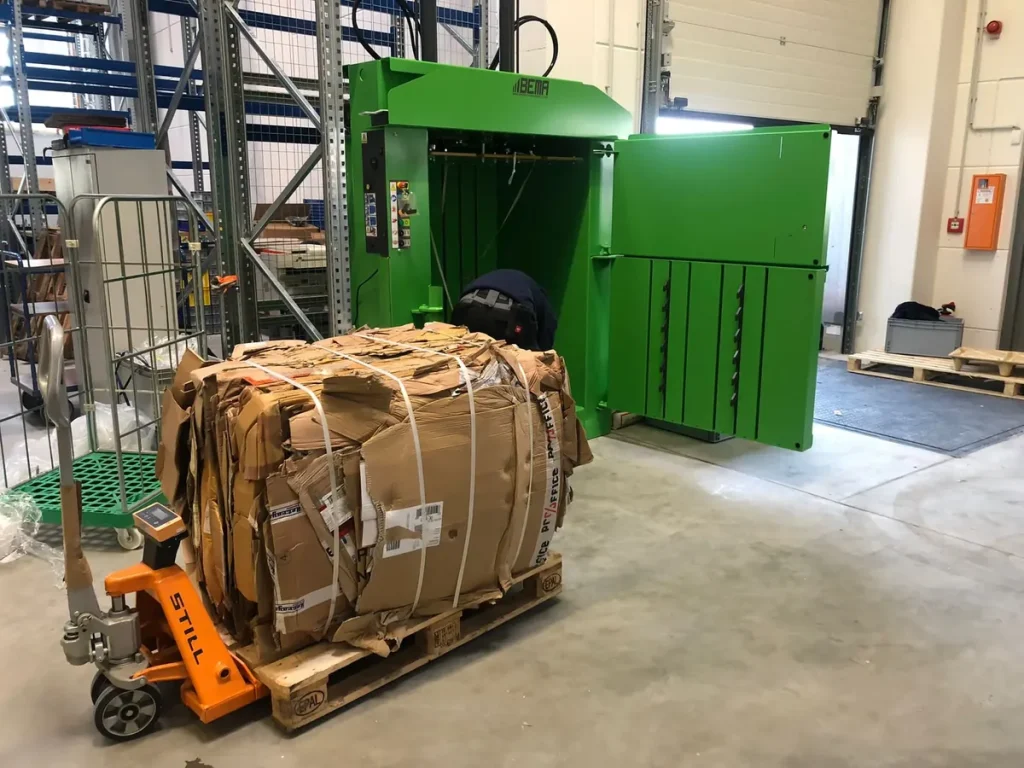 Gepresster Kartonballen auf einem Hubwagen vor einer geöffneten BEMA Ballenpresse
