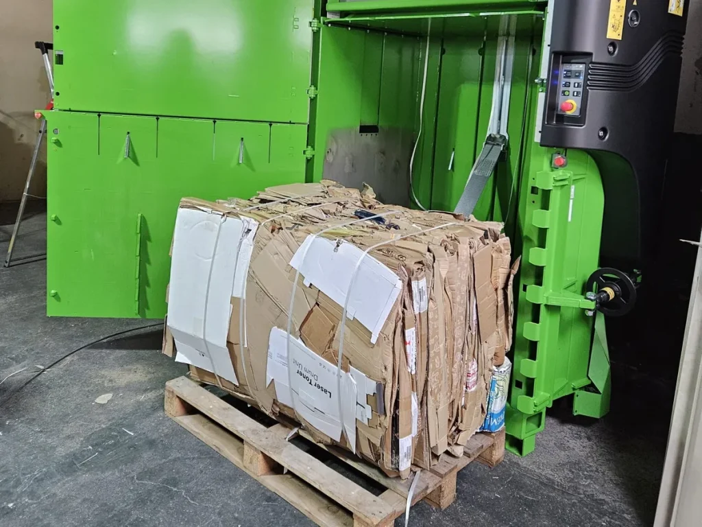 Gepresster Kartonballen auf einer Palette vor einer geöffneten Ballenpresse